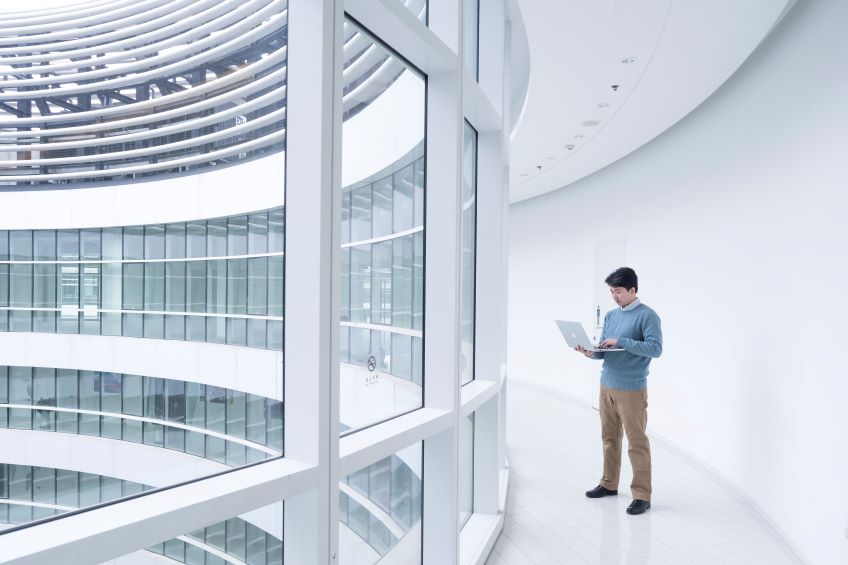 An individual stands in a modern, spacious atrium with a white interior, holding and looking at an open laptop, with a curved, layered ceiling design allowing natural light to filter through