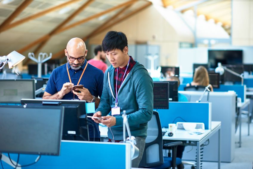 An indoor office environment with multiple workstations, where individuals are seated at desks with computer monitors, and two individuals are standing in the foreground, engaged in a conversation