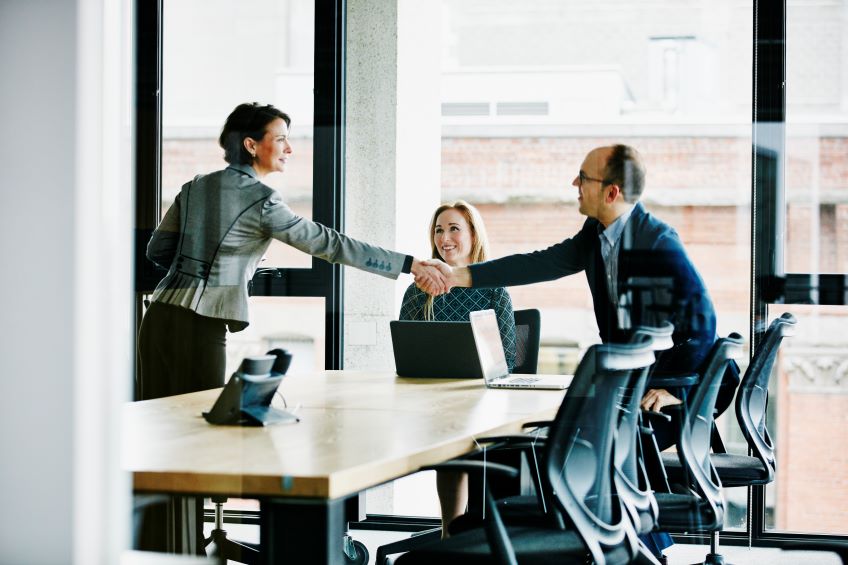 Two individuals shaking hands across a table, with a laptop open in front of them and another person seated at the table in a conference room