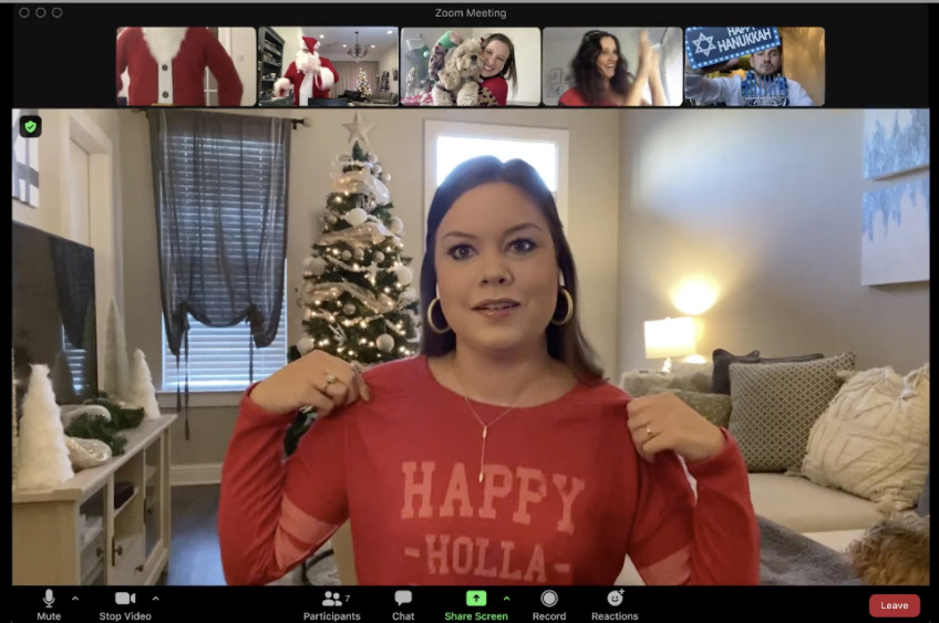 A screenshot of a Zoom meeting with multiple participants, focusing on one participant in a red sweater with the text "HAPPY HOLLA-DAYS," standing in front of a Christmas tree and decorations