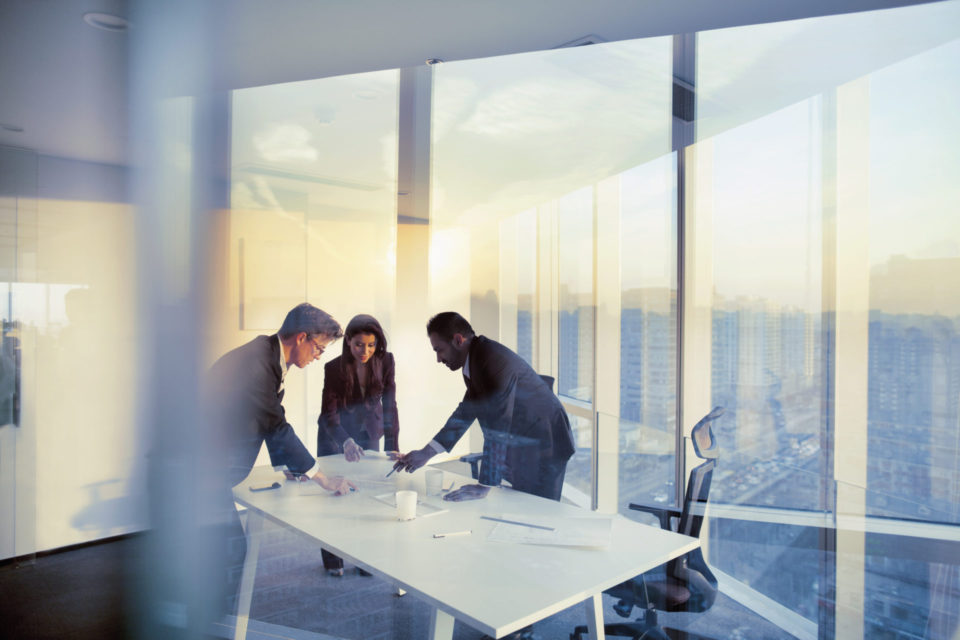 Three individuals in a modern office setting with large windows offering a view of the city skyline during sunset, engaged in a discussion over documents on a table