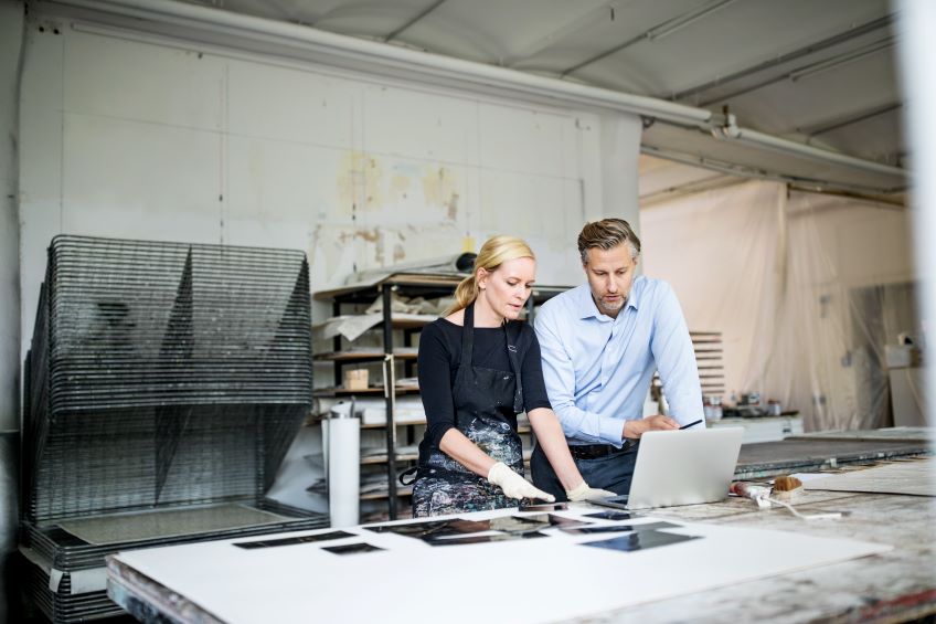 Two individuals stand in front of an open laptop in a studio environment, with shelves in the background