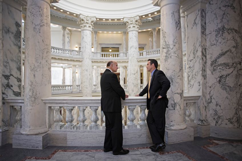 Two individuals stand in a grand interior space with columns, balustrades and marble surfaces