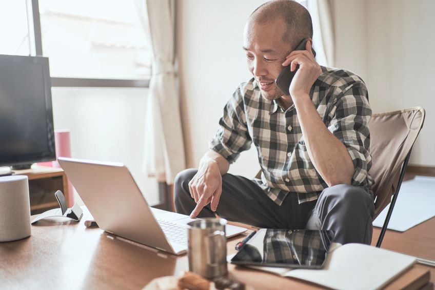 An individual sitting at a desk, interacting with a laptop and speaking on a mobile phone in a home office environment