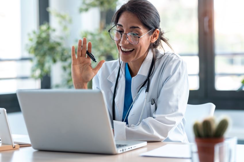 A person in a medical coat sits before a laptop, smiling and waving into the camera