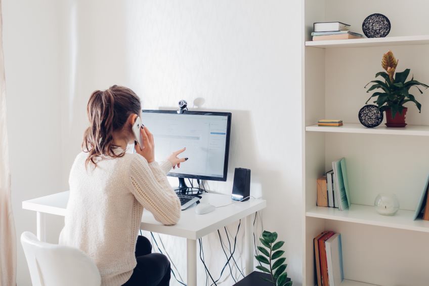 An individual sitting at a desk in front of a computer monitor, pointing at the screen with the right hand while holding a smartphone to the ear with the left hand