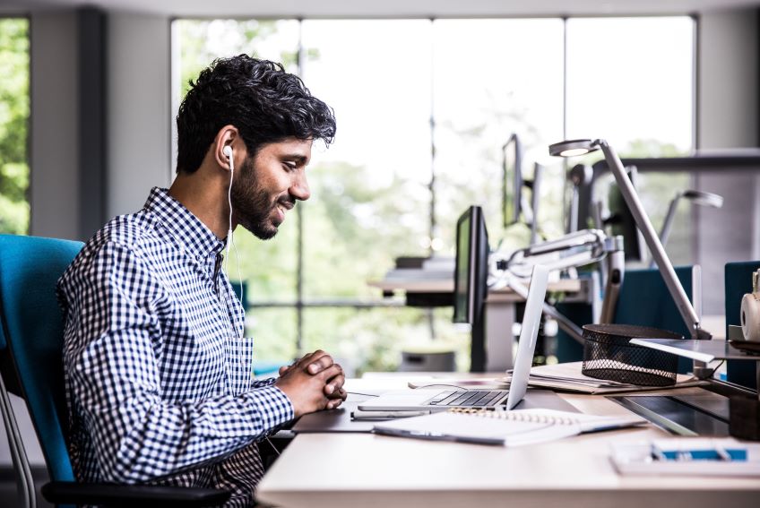 A person in a modern office sits at a desk in front of a window with headphones viewing or listening to a conference call on Zoom