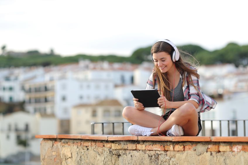 A person sitting outside on a brick wall watching a tablet with headphones with houses in the background