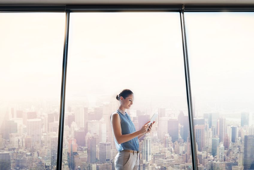 Woman stands with tablet In front of large windows with cityscape background