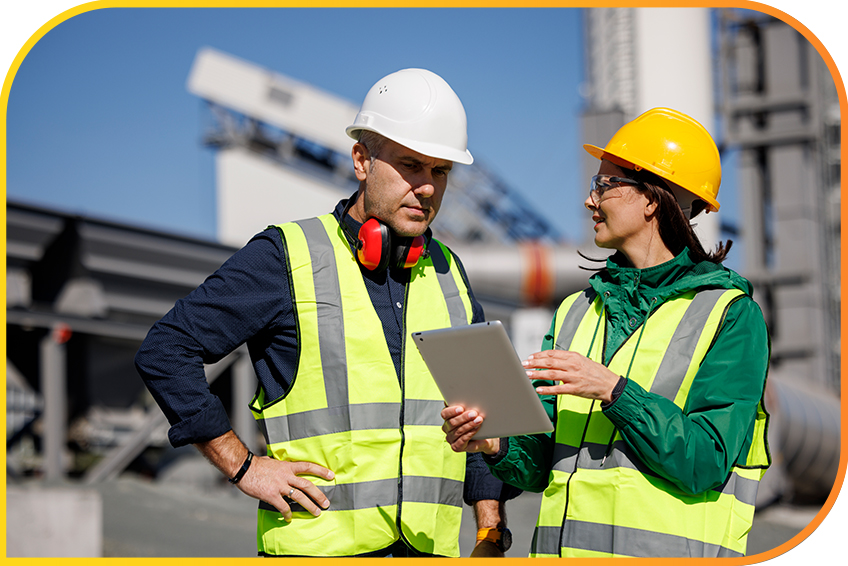 Two people wearing safety vests and helmets have a discussion while looking at a tablet.