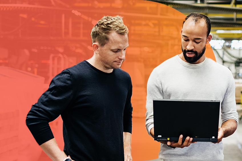 Two professionals having a conversation stand side by side with one holding a laptop in a grey sweater and another with hands on hips in a black sweater