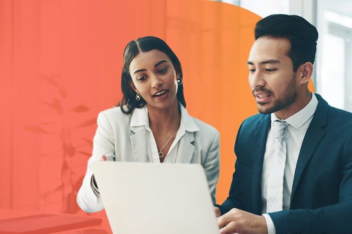 Two people in formal business attire talk while in front of a laptop over an orange background